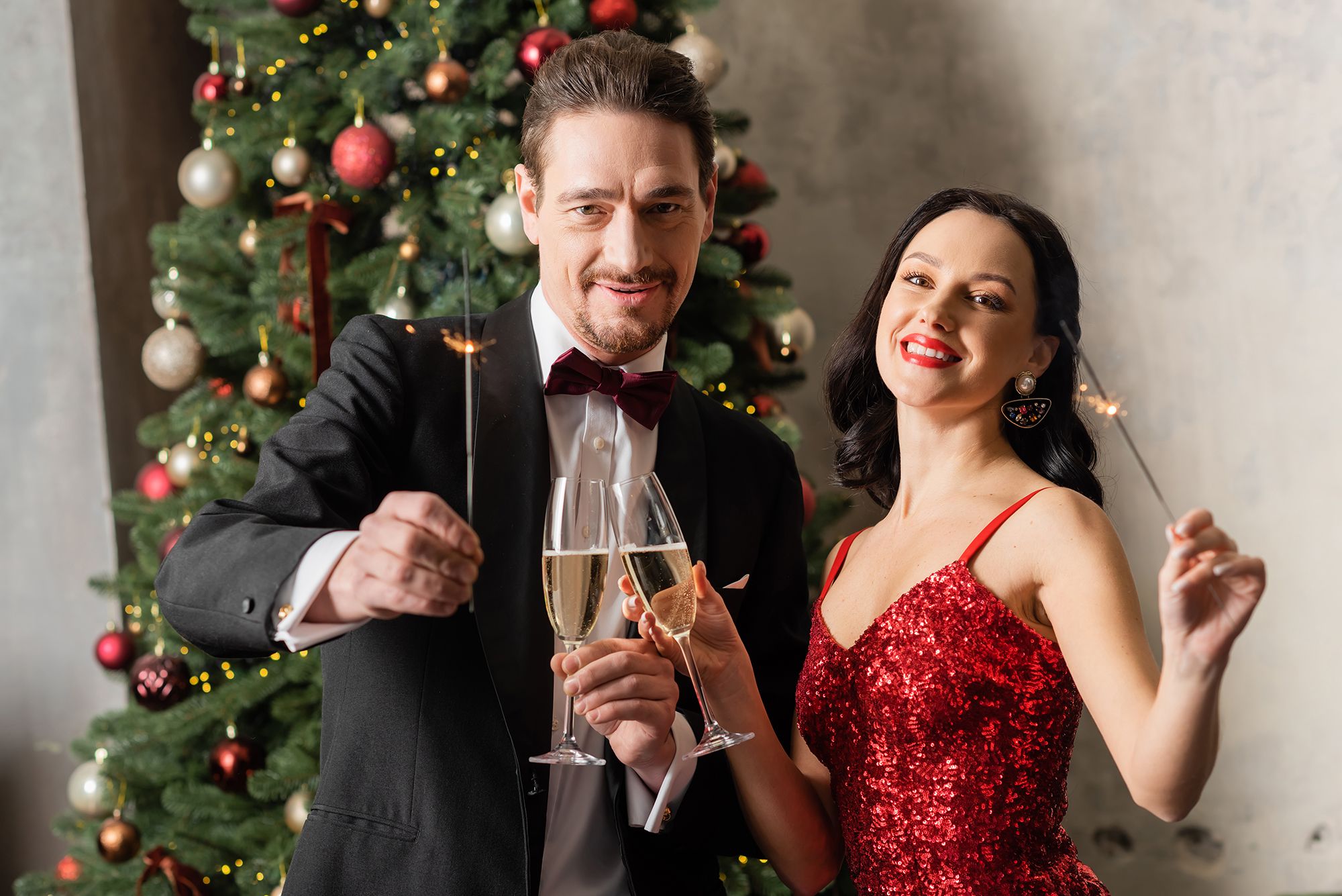 A happy couple celebrates Christmas out in Miami, she is wearing a red dress and him a tux and they are toasting with champagne and a Christmas tree in the background while holding sparklers.